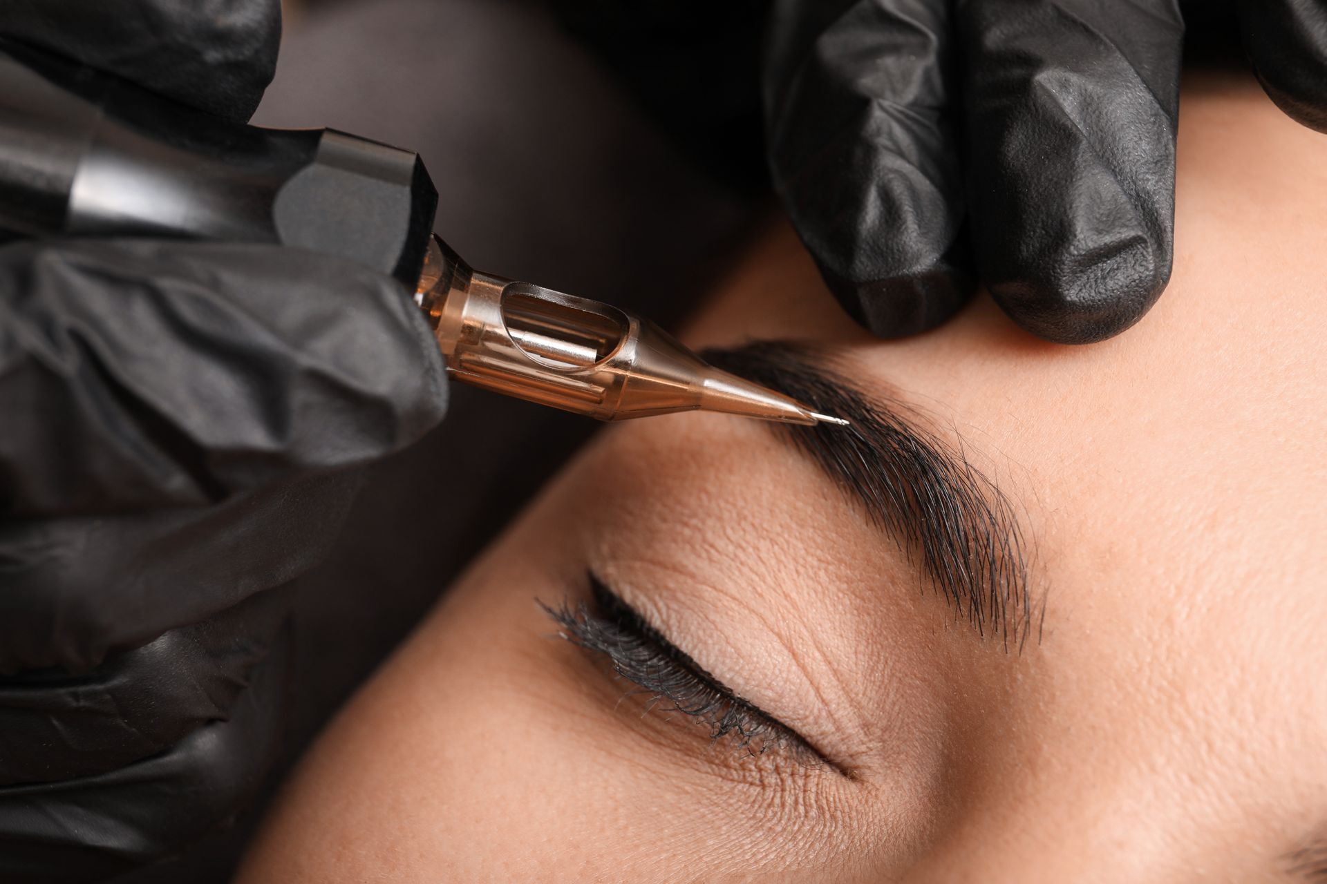 Microblading der Augenbrauen in Arbeit: Behandschuhte Hände halten ein Gerät in der Nähe der Augenbraue einer Frau.