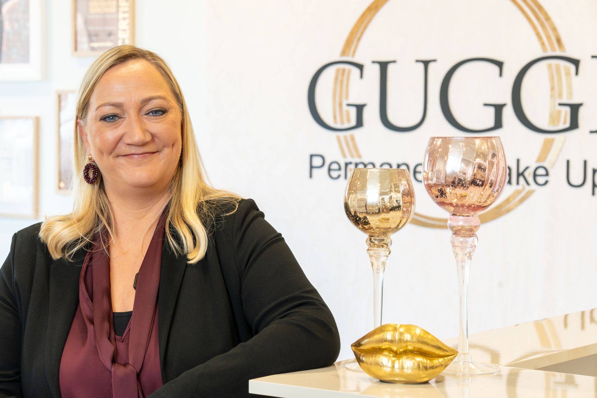 Eine Frau steht vor einem Logo mit dekorativen Kerzen. Sie lächelt, trägt einen Blazer und steht in einem Innenraum.