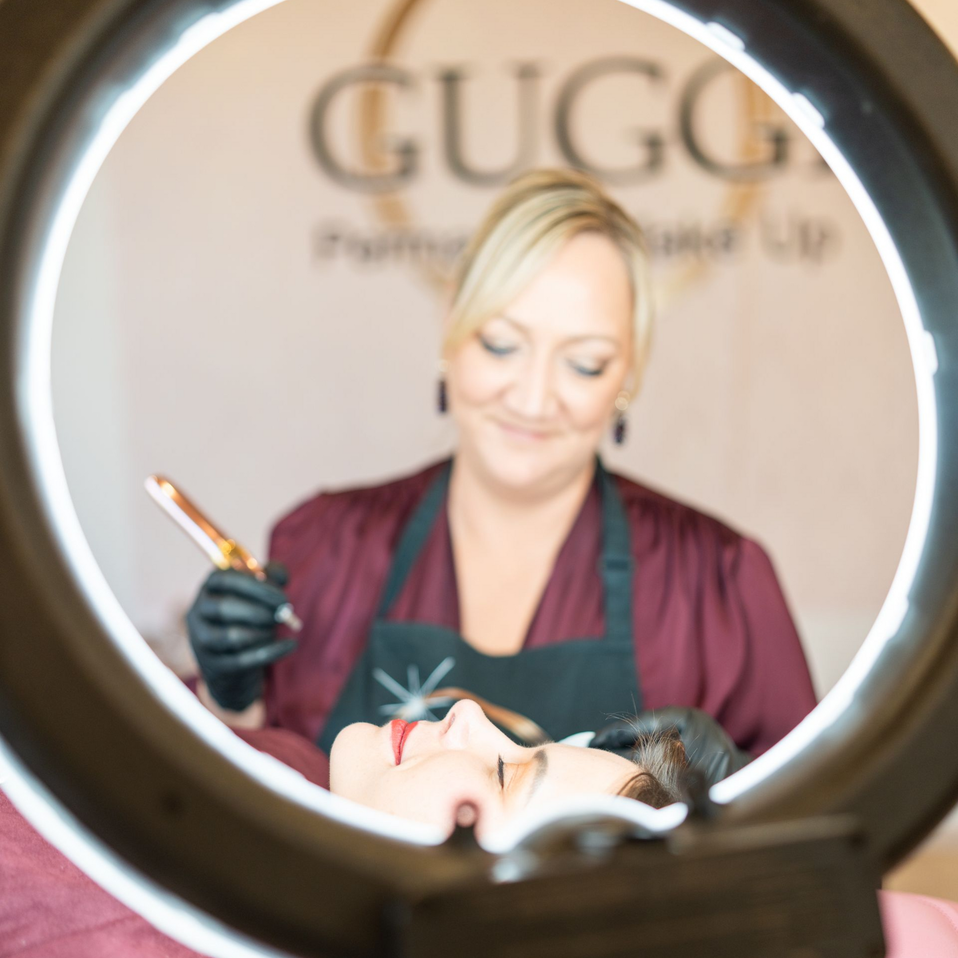 Eine Frau führt bei einer Kundin eine Permanent-Make-up-Behandlung durch, die von einem Ringlicht beleuchtet wird.