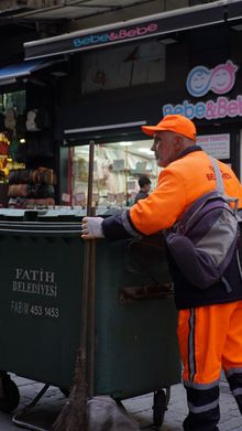 Orange-clad sanitation worker pushing a green trash bin past a storefront with neon signage