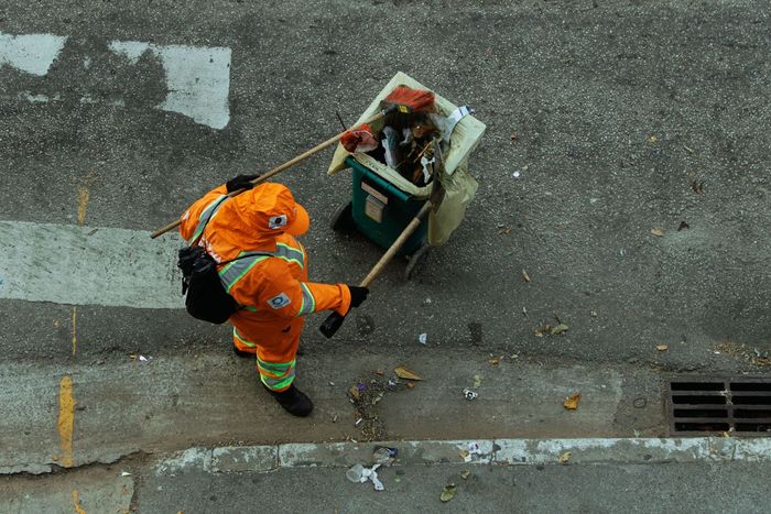 Two road workers in orange uniforms push a trash cart along a curb