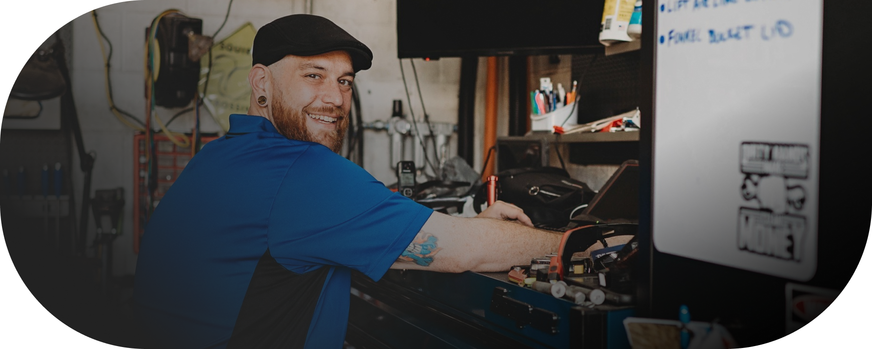 Man wearing a blue shirt and a black cap smiles while working at a workbench in a workshop | City Side Auto