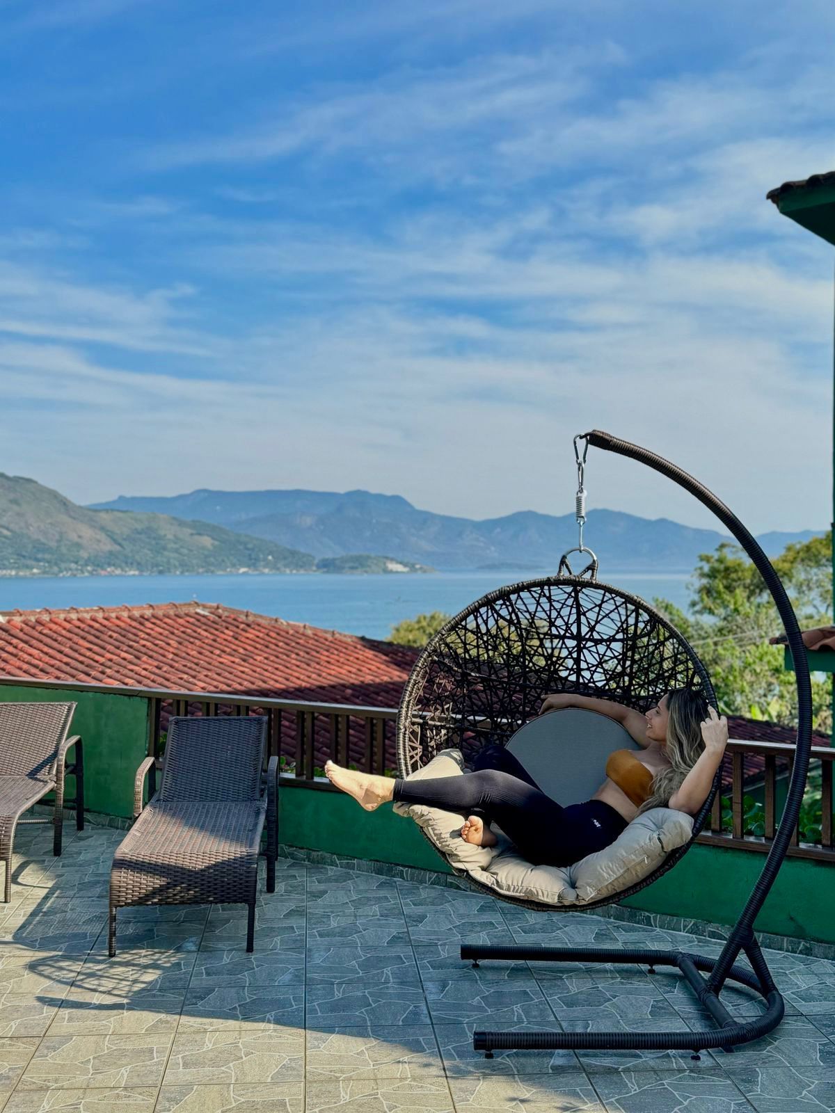 Mulher relaxando em uma cadeira suspensa em um pátio com vista para um lago e montanhas sob um céu azul.