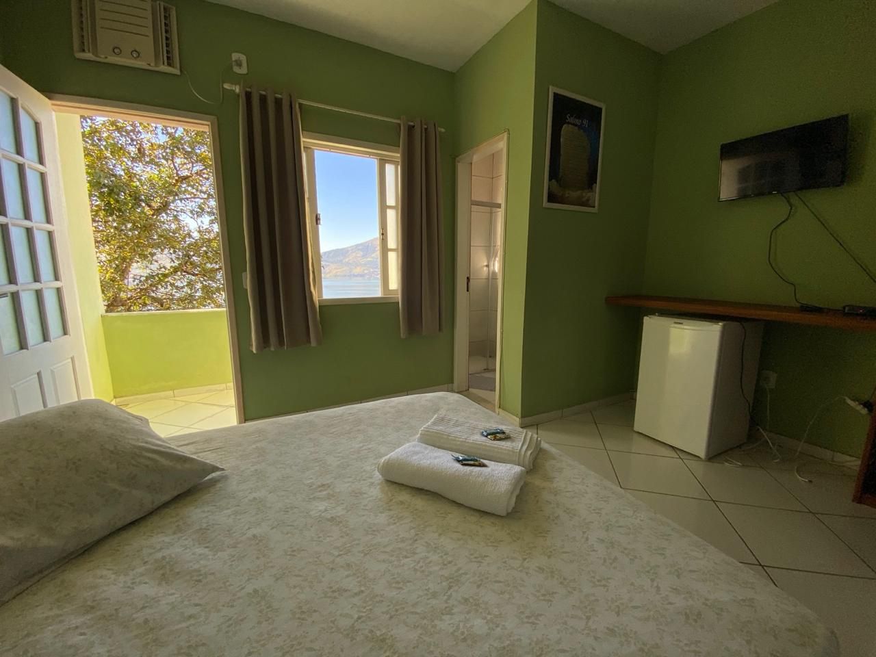 Quarto com paredes verdes, cama, frigobar, janela com vista para o mar e porta aberta para uma varanda.