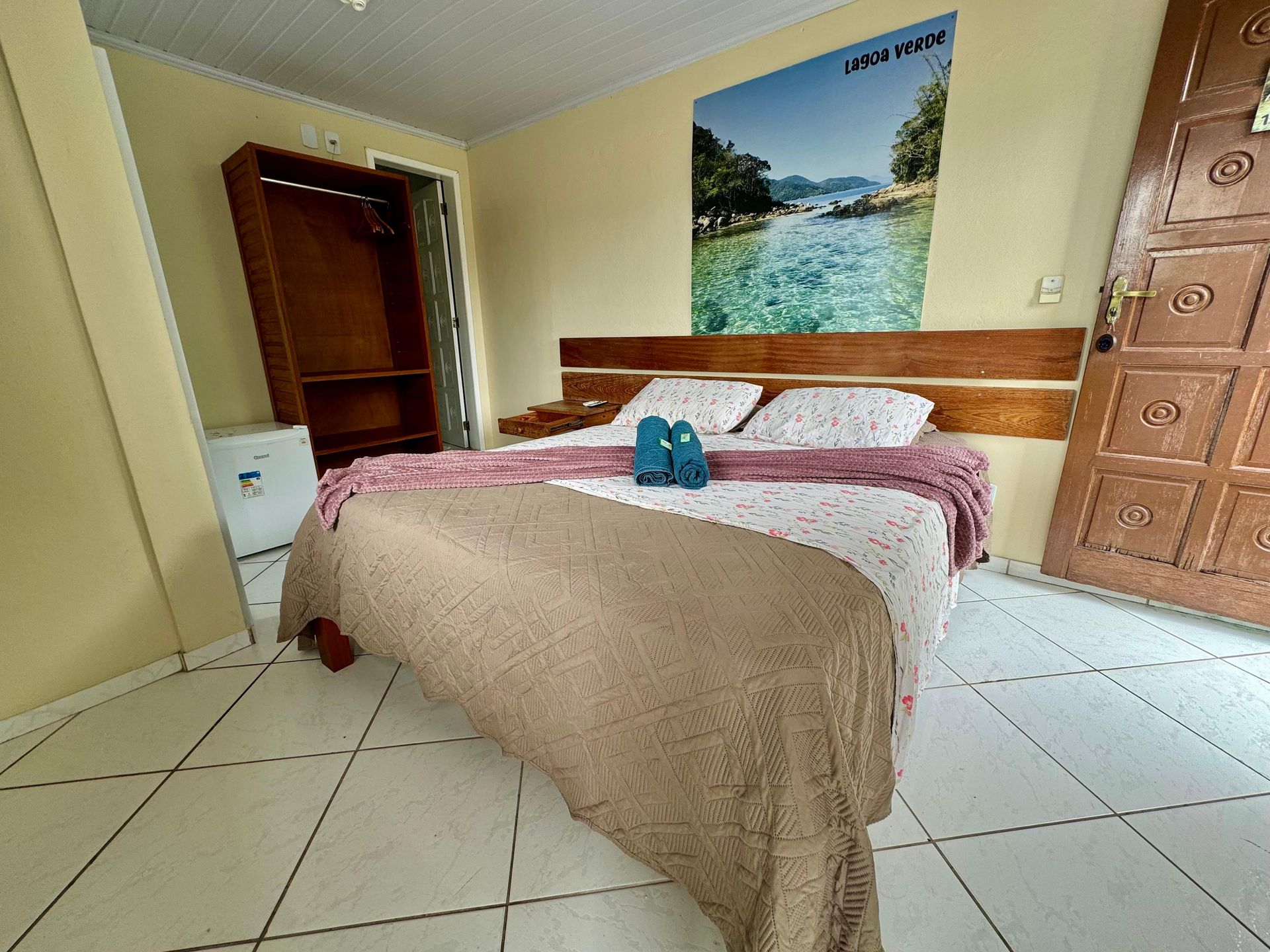 Quarto com cama de casal, guarda-roupa, frigobar e porta. Quadro com paisagem fluvial na parede.