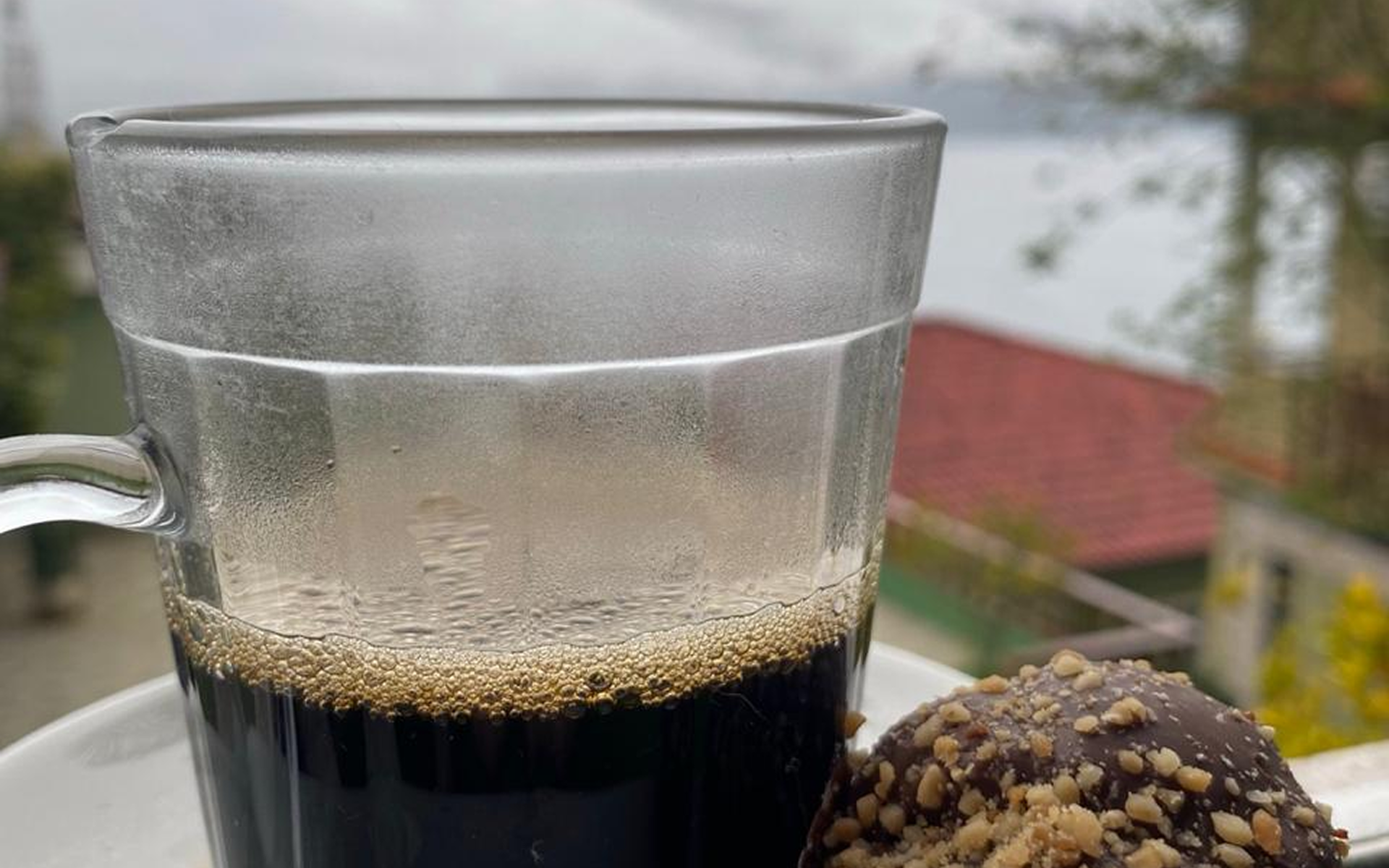 Caneca de vidro transparente com café escuro sobre um pires, com um doce coberto de chocolate ao lado, em um ambiente externo.