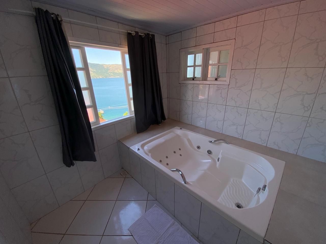 Banheiro com banheira de hidromassagem. As janelas oferecem uma vista panorâmica do oceano; cortinas cinza. Azulejos brancos.