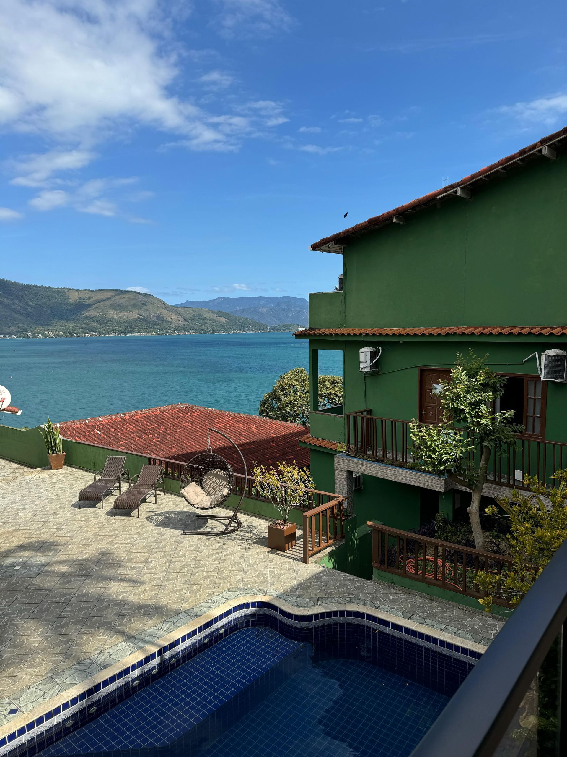 Prédio verde à beira-mar azul com piscina e espreguiçadeiras. Telhado marrom e céu limpo à vista.
