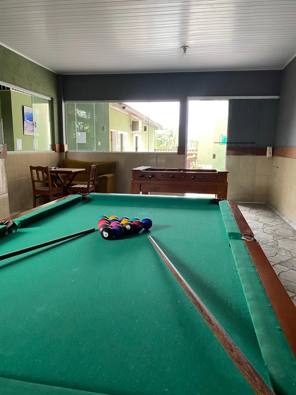 Mesa de bilhar com bolas enfileiradas, tacos, em uma sala com janela, cadeiras e sofá.