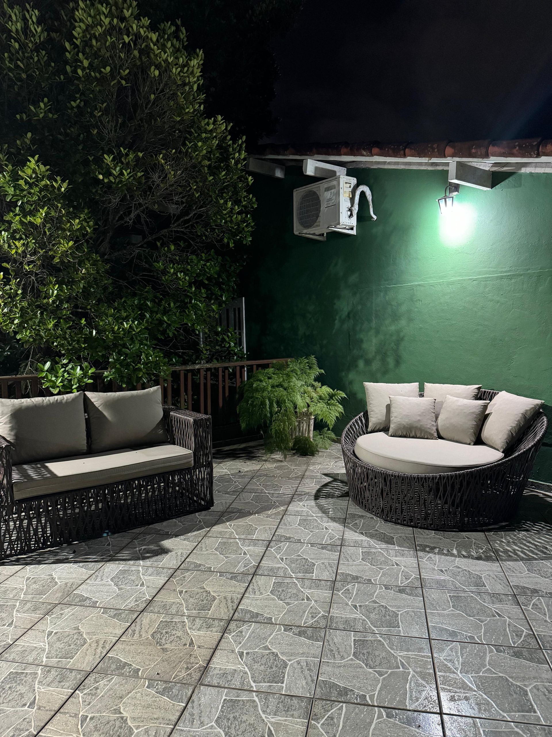 Mobiliário de jardim em um pátio de pedra à noite. Parede verde, árvores e aparelho de ar condicionado.