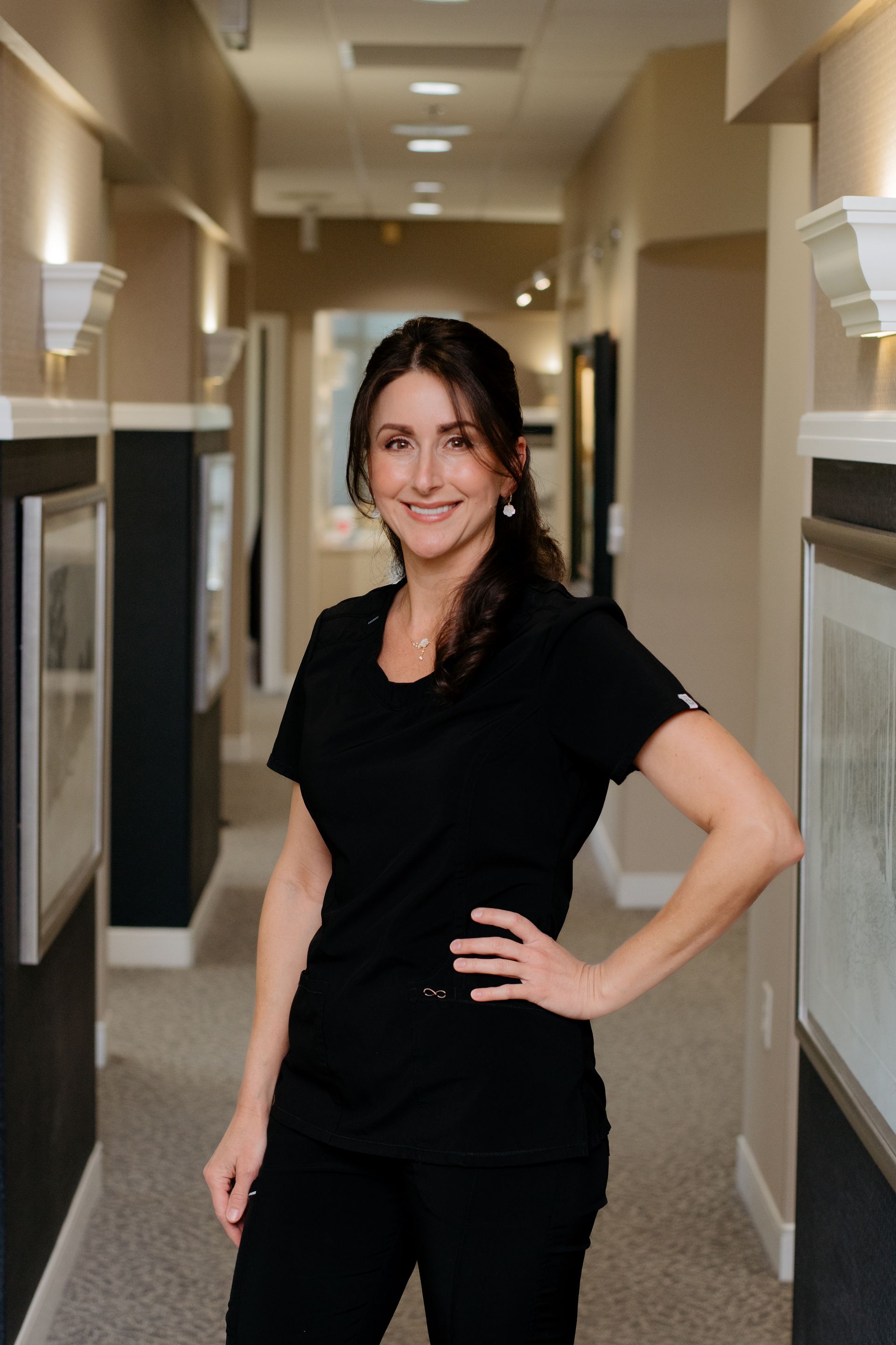 A woman in a black scrub top is standing in a hallway and smiling.