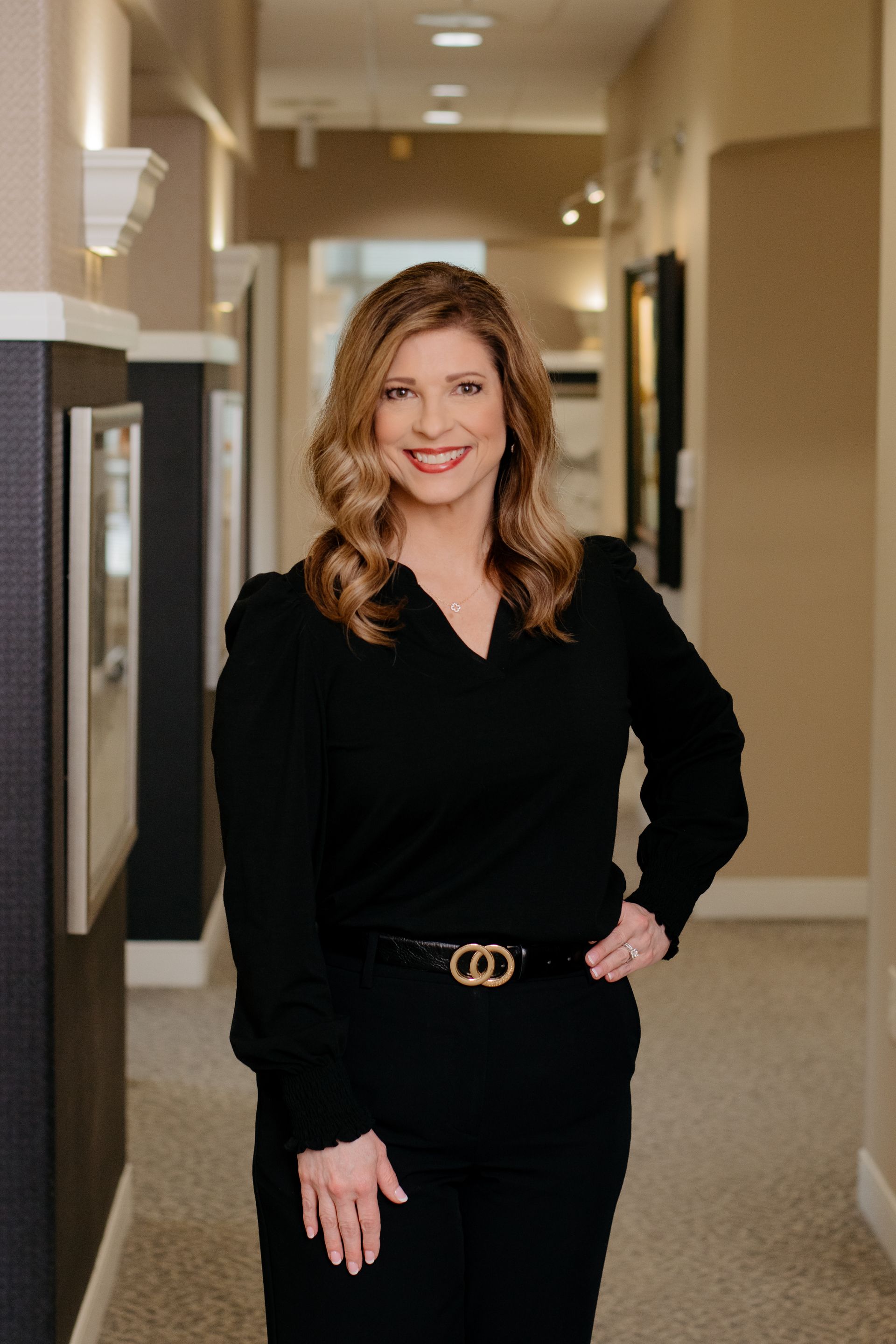 A woman is standing in a hallway with her hands folded and smiling.
