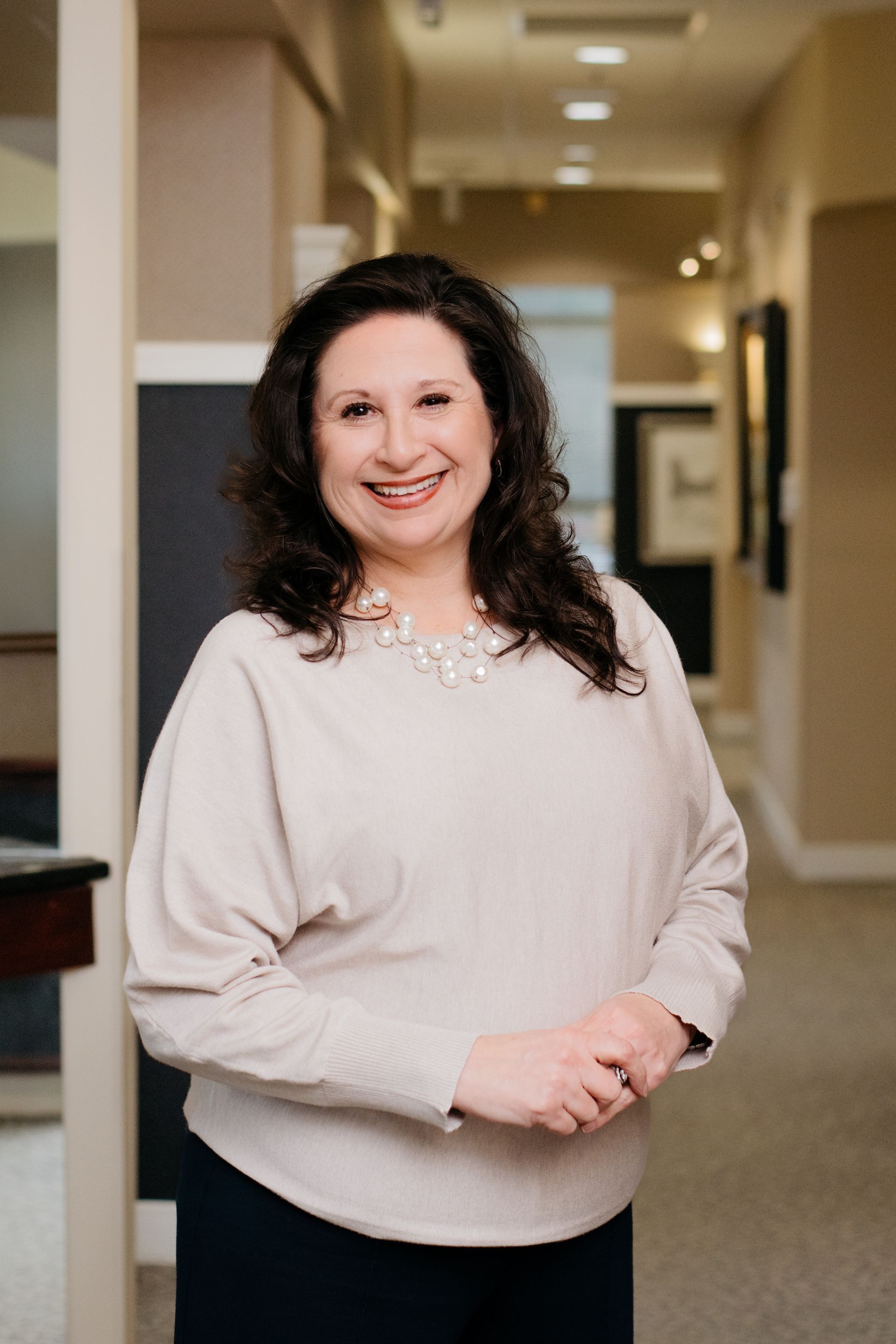 A woman is standing in a hallway with her hands folded and smiling.