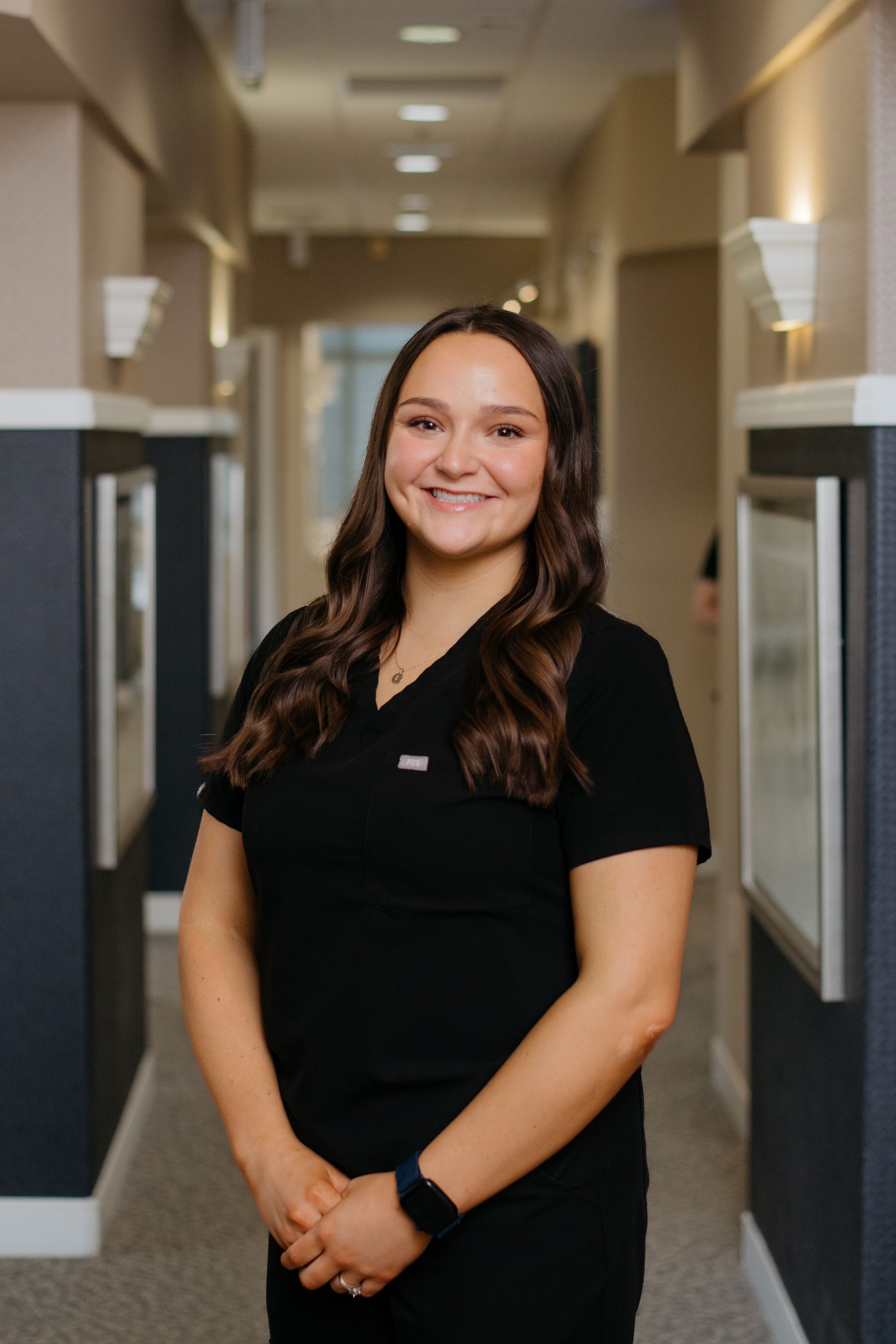 A woman in a black scrub top is standing in a hallway and smiling.