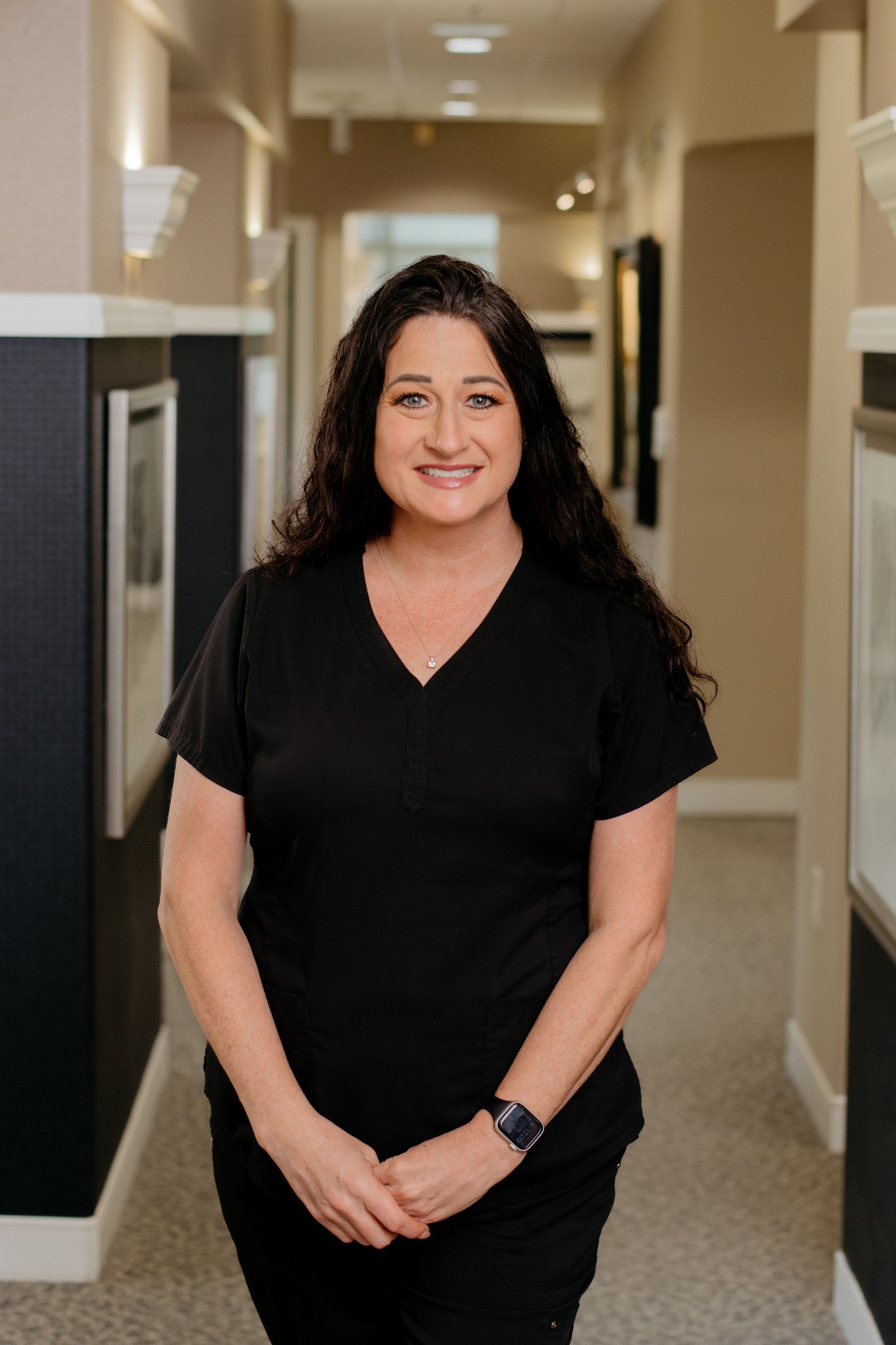 A woman in a black scrub top is standing in a hallway with her arms crossed.