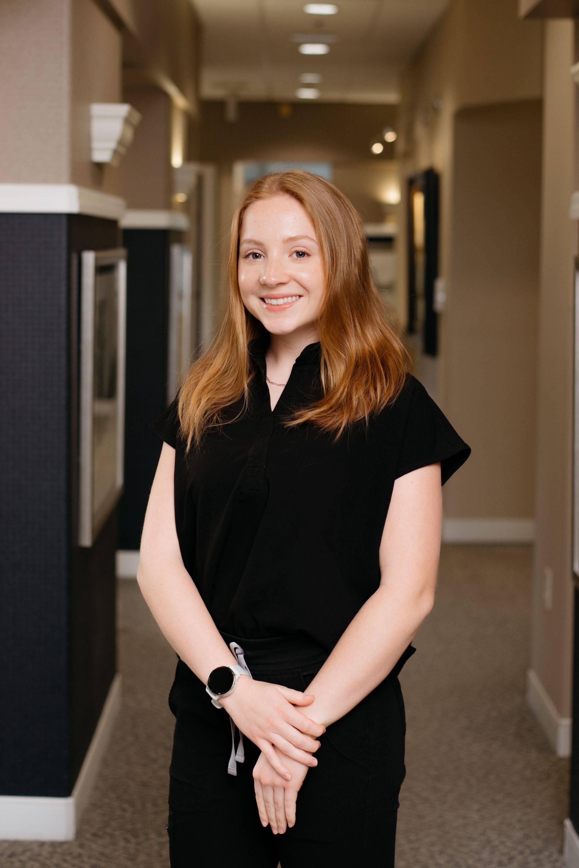 A woman is standing in a hallway with her hands folded and smiling.