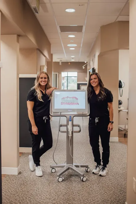 Two women in scrubs are standing next to a machine in a hallway.