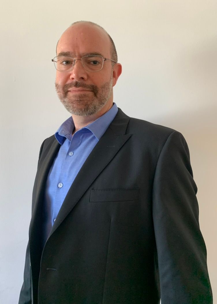 A man in a suit and blue shirt is standing in front of a white wall