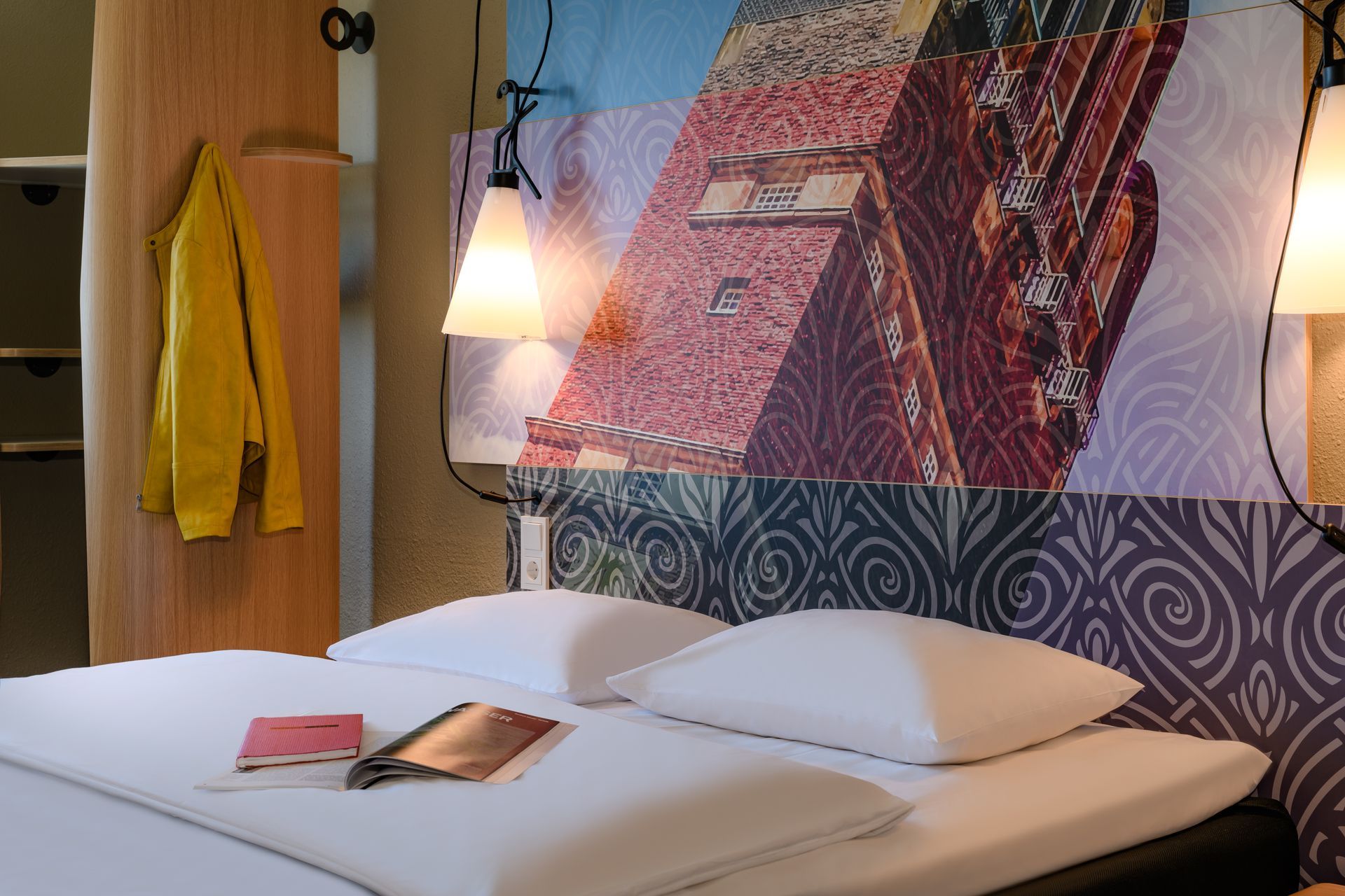 Modern hotel room with made bed, hanging lamp, colorful wall art, and towels on the wall
