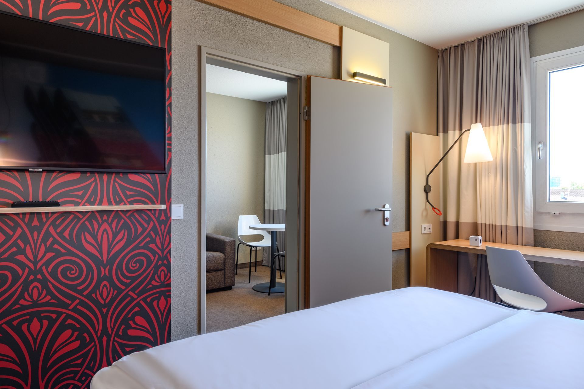 Modern hotel room with bed, desk, TV, mirror, and window lighting