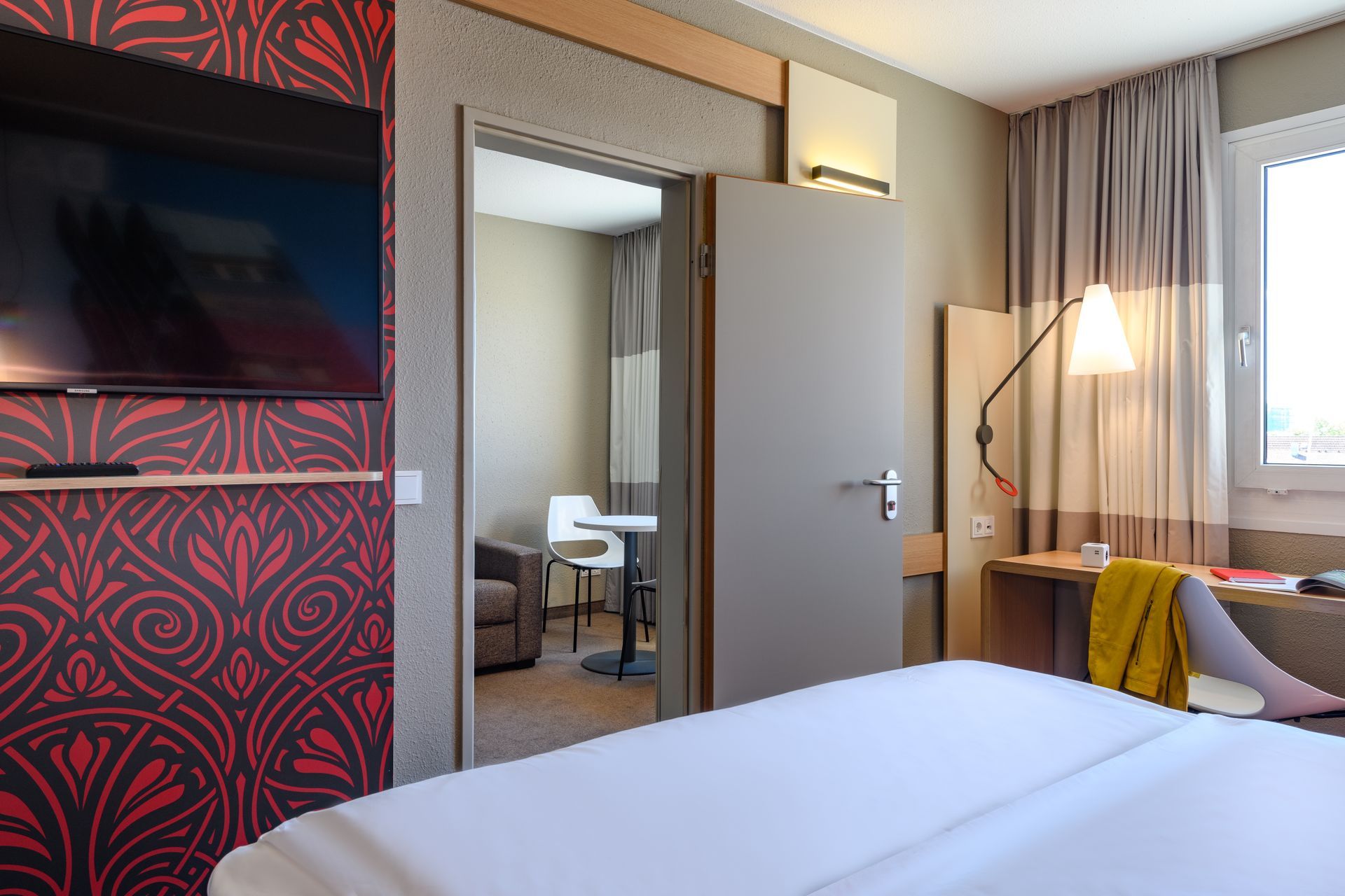 Modernes Hotelzimmer mit Bett, Wandfernseher, Spiegel, Schreibtischlampe und Fenstervorhängen.