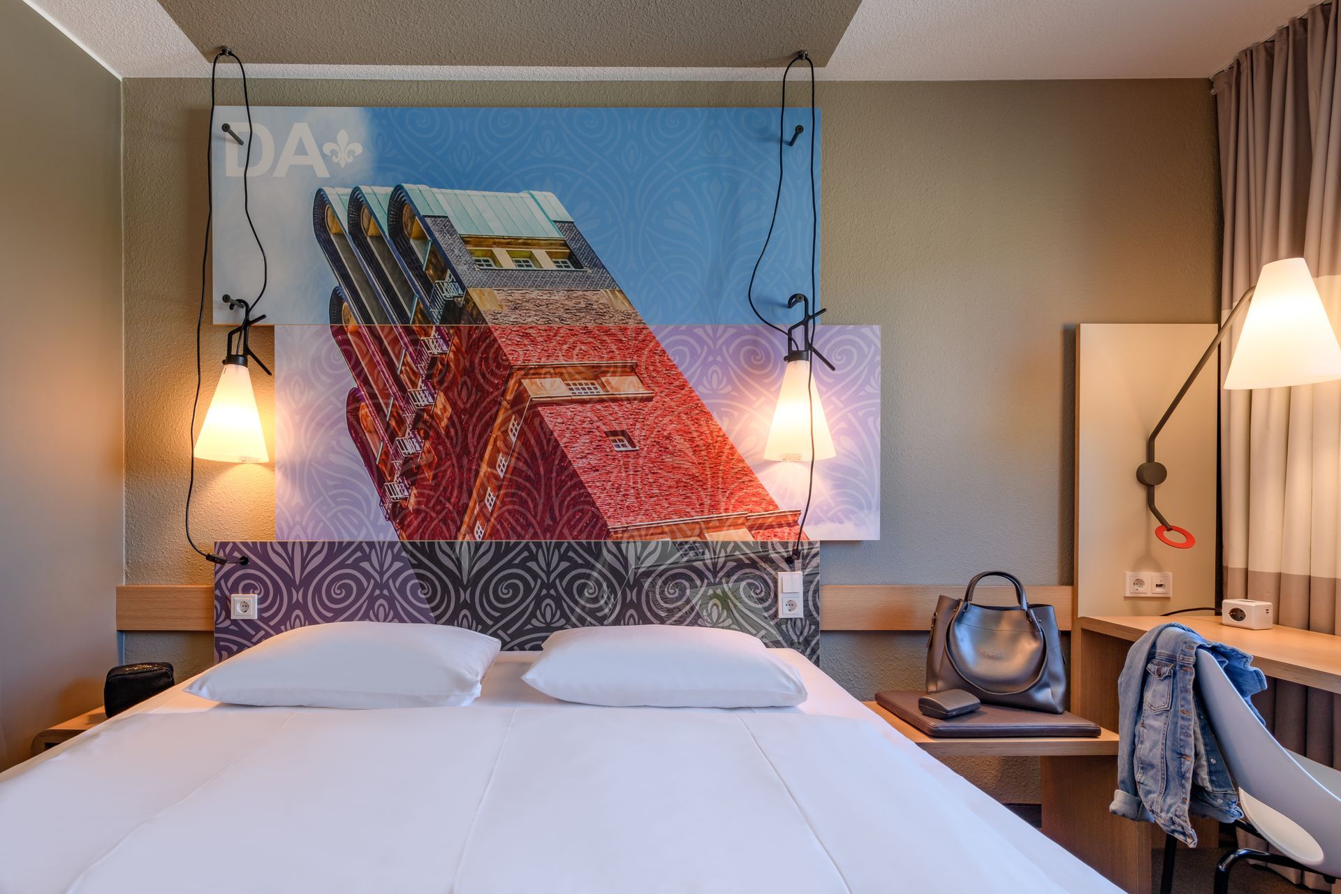 Hotelzimmer mit Bett, farbenfrohem Kopfteilbild, Pendelleuchten und einem Schreibtisch am Fenster