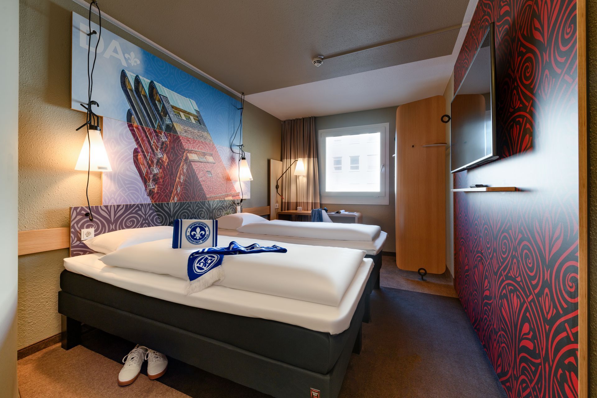 Hotelzimmer mit zwei Betten, gemusterten Wandbildern und einem Fenster am Ende des Zimmers