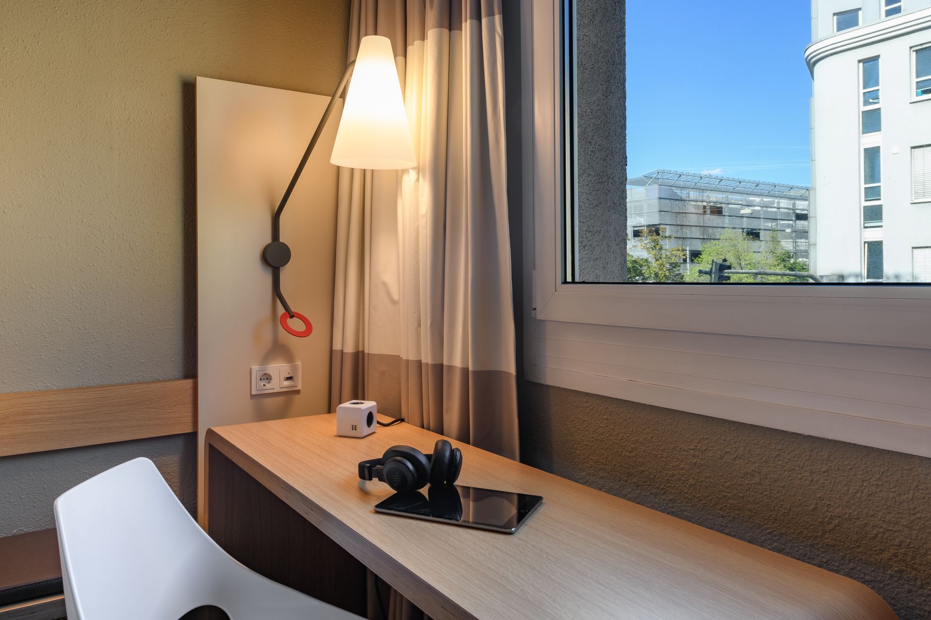 Hotel desk by window with lamp, curtains, chair, camera, and city view