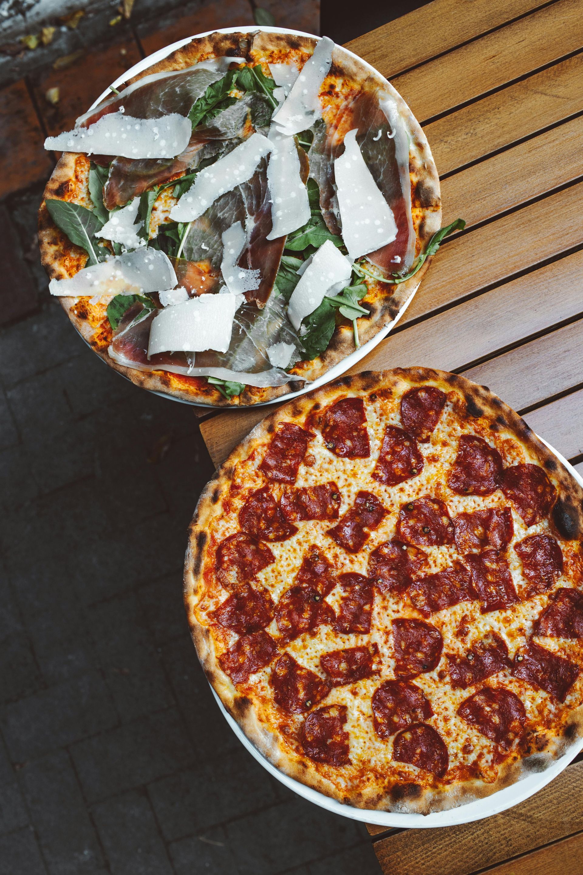 Two pizzas on a wooden table: one topped with greens and white strips, one pepperoni and cheese.