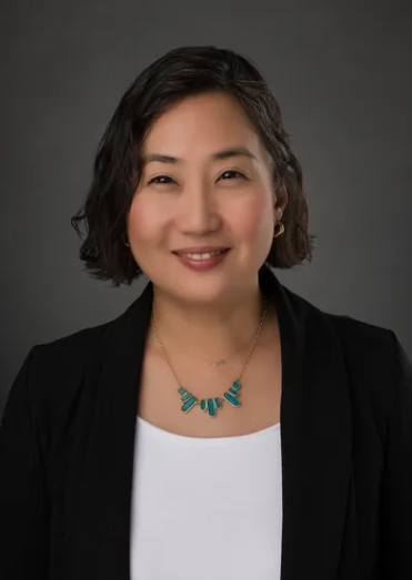 A woman wearing a black jacket and a necklace is smiling for the camera.