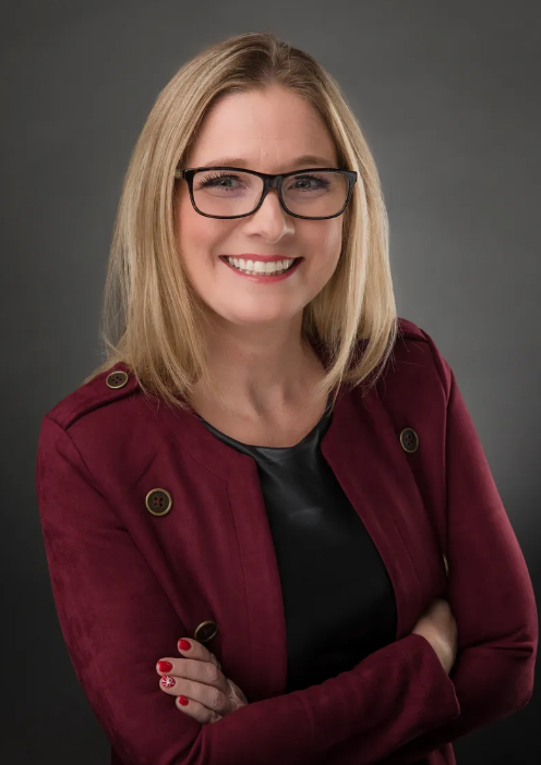 A woman wearing glasses and a red jacket is smiling with her arms crossed.