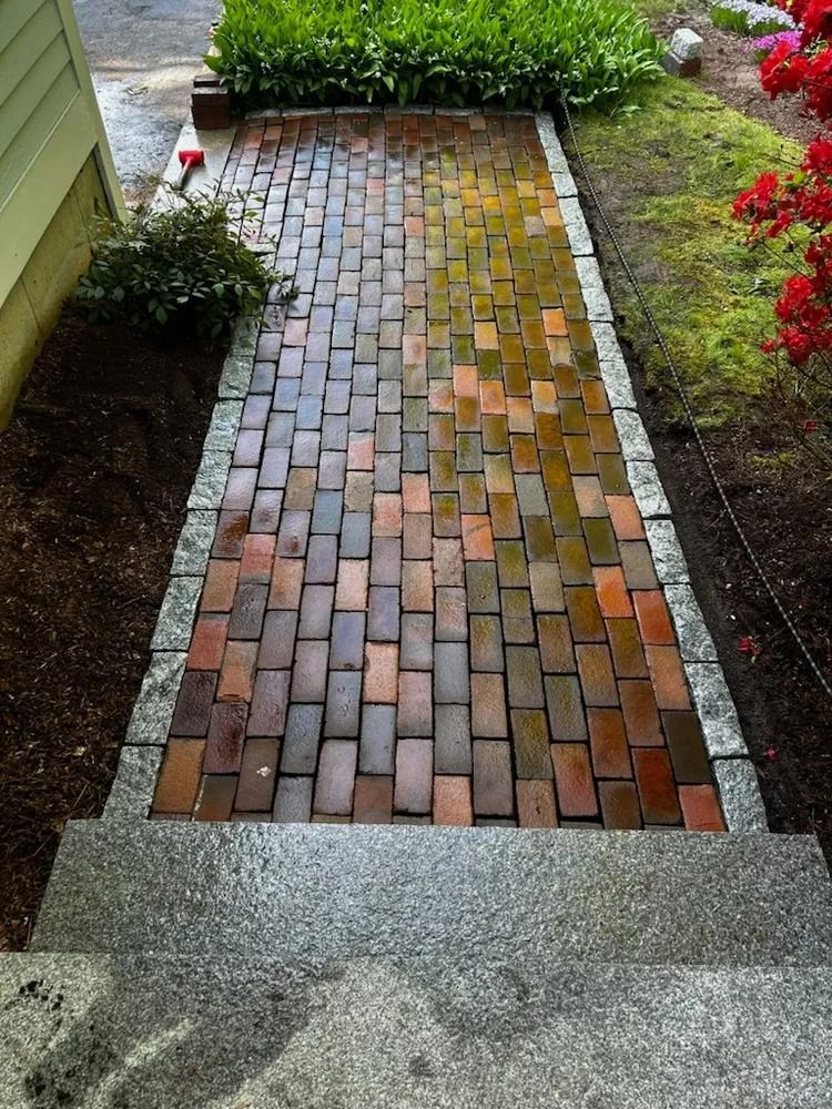 Brick walkway bordered by gravel, leading from steps up to a garden, with green plants and red flowers.