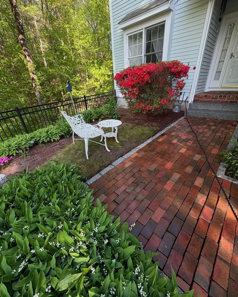 Brick path leads to a house with red azalea bushes, white bench, and table. Green foliage surrounds the scene.