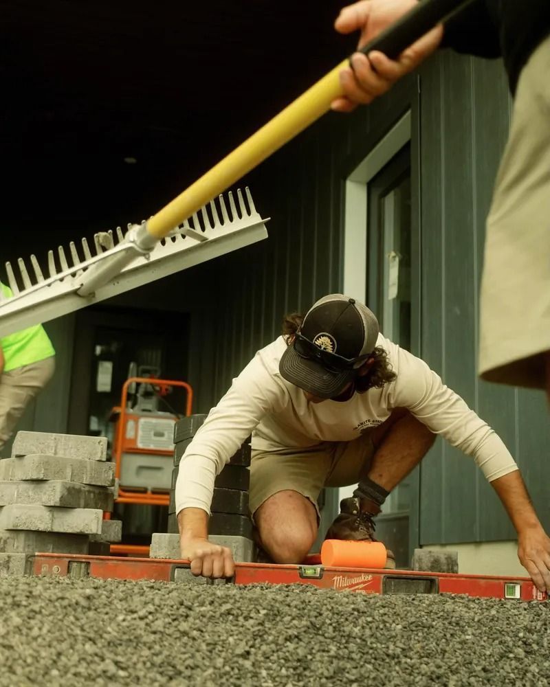 Man kneeling, laying pavers; another holds rake. Construction site with gravel and tools.