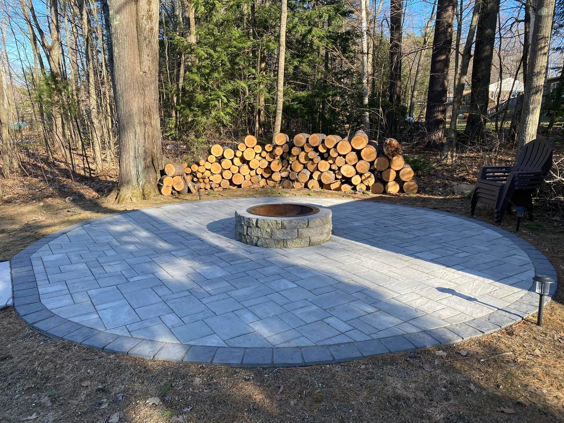 Circular paved patio with fire pit, firewood, and a lawn chair surrounded by trees.