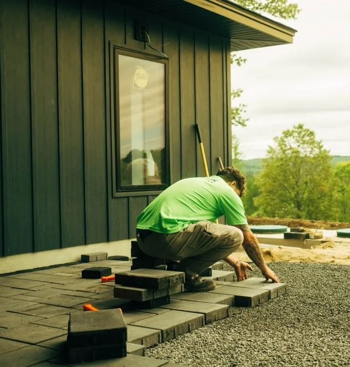 Man laying pavers on a patio near a dark-sided building; gravel and trees in background.