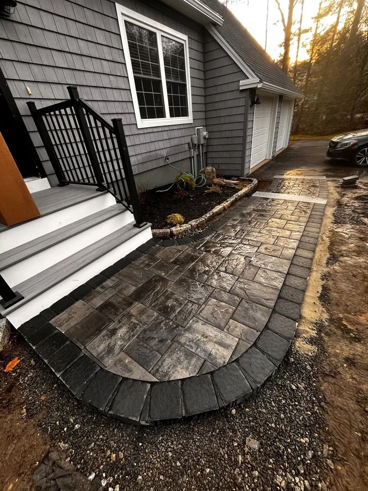 Pathway with brick-like pavers leading to gray house with black railing. Black border surrounds the pathway.