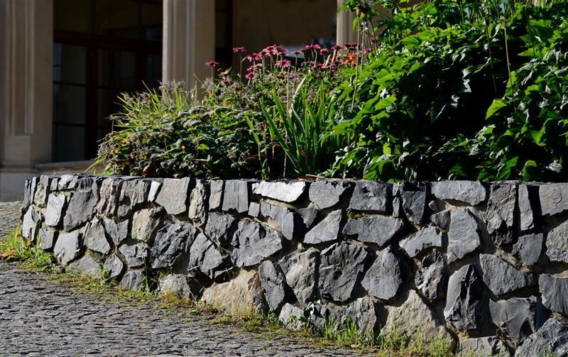 Stone retaining wall with lush green plants and flowers in front of a building.