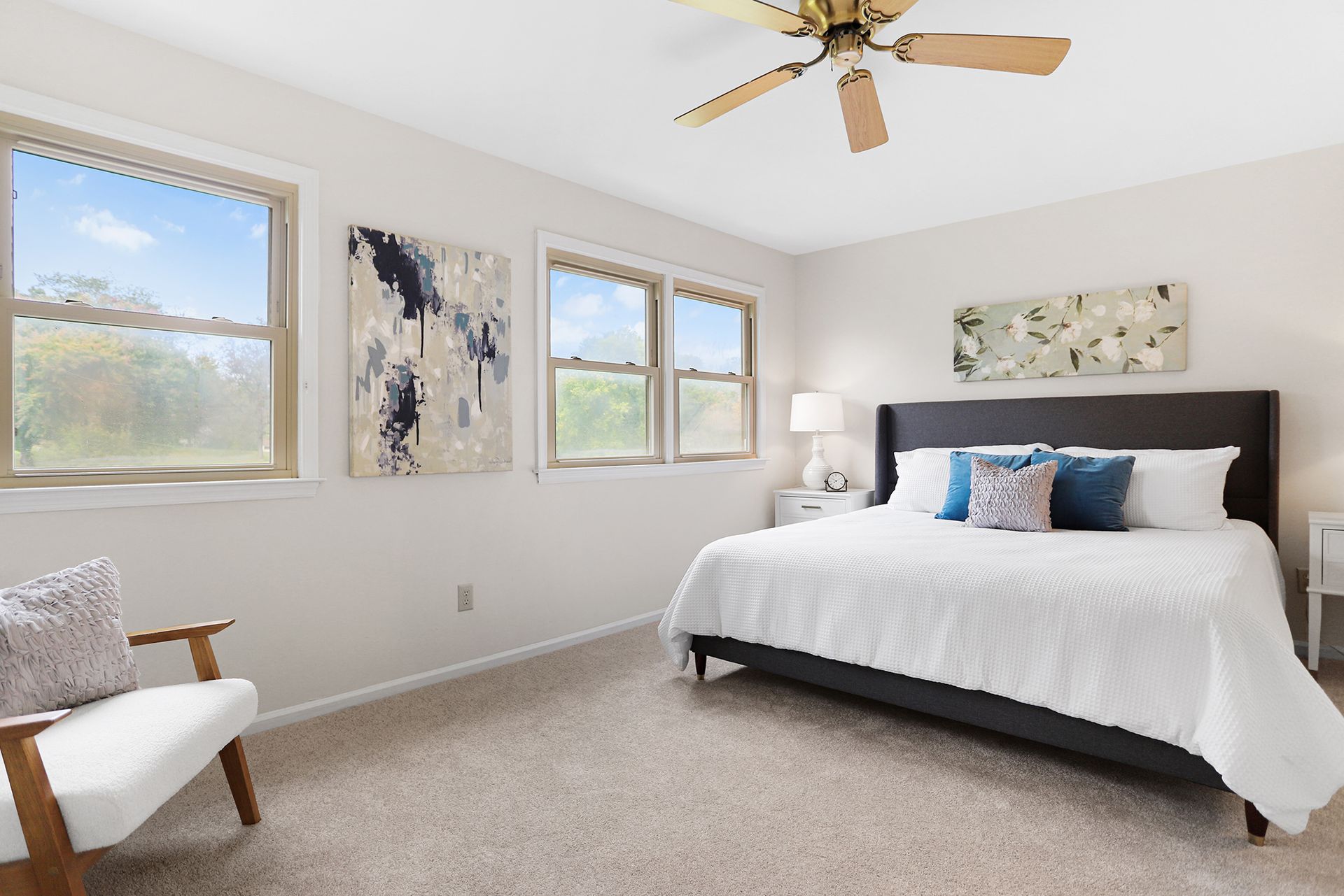Bedroom with a bed, two nightstands, a chair, and three windows; neutral colors.