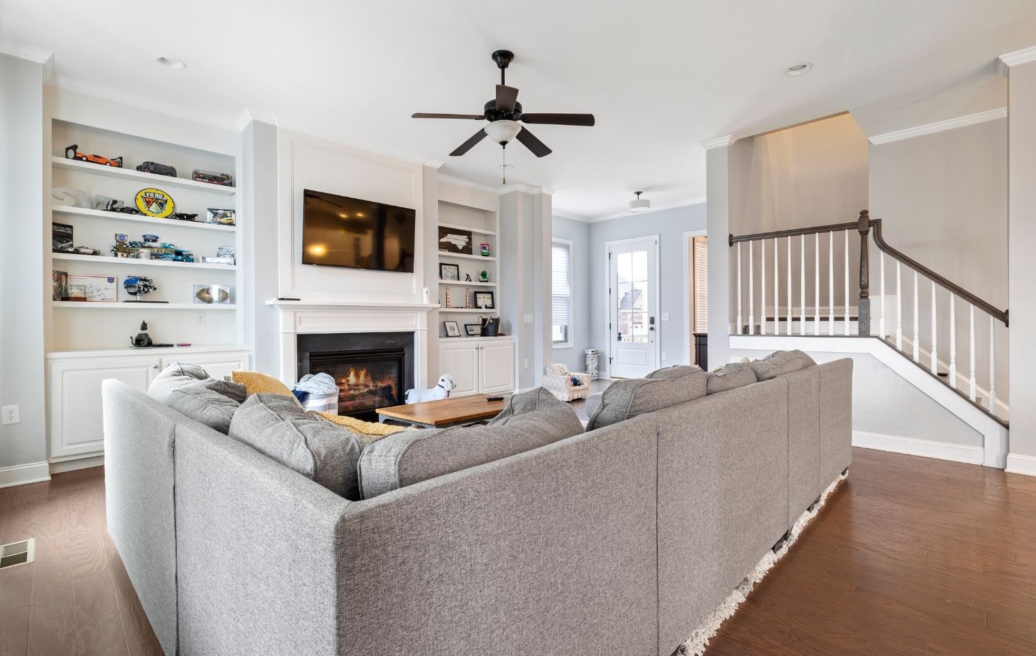 Spacious living room with gray sectional sofa, fireplace, built-in shelves, and staircase.