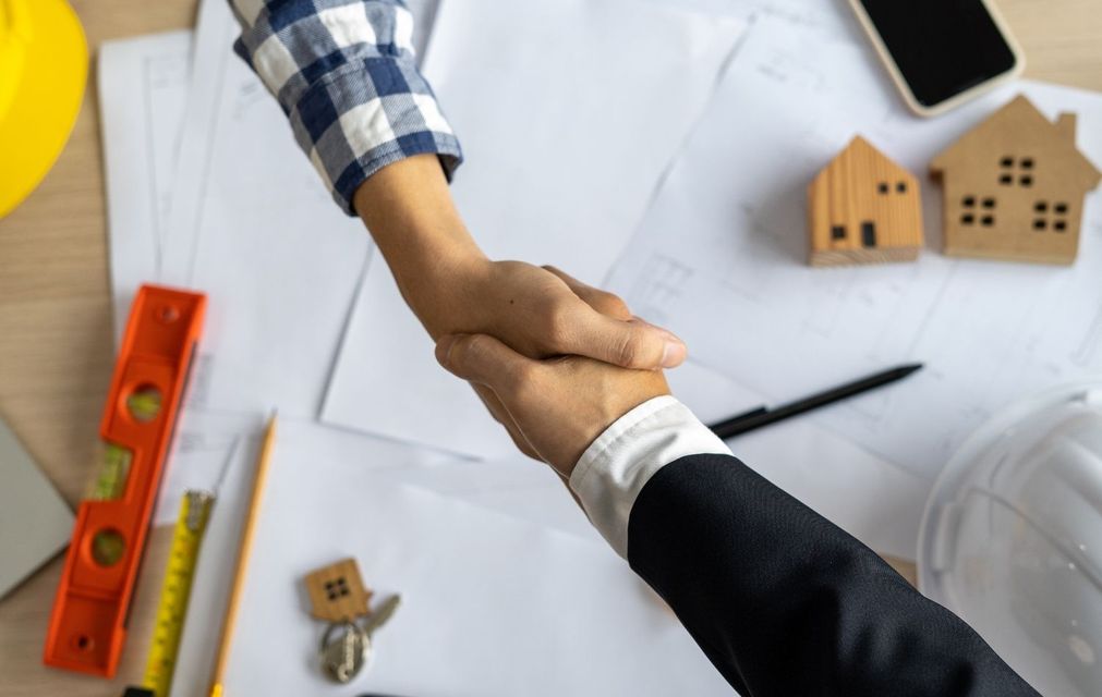 Two people shaking hands over blueprints and miniature house models, symbolizing an agreement.