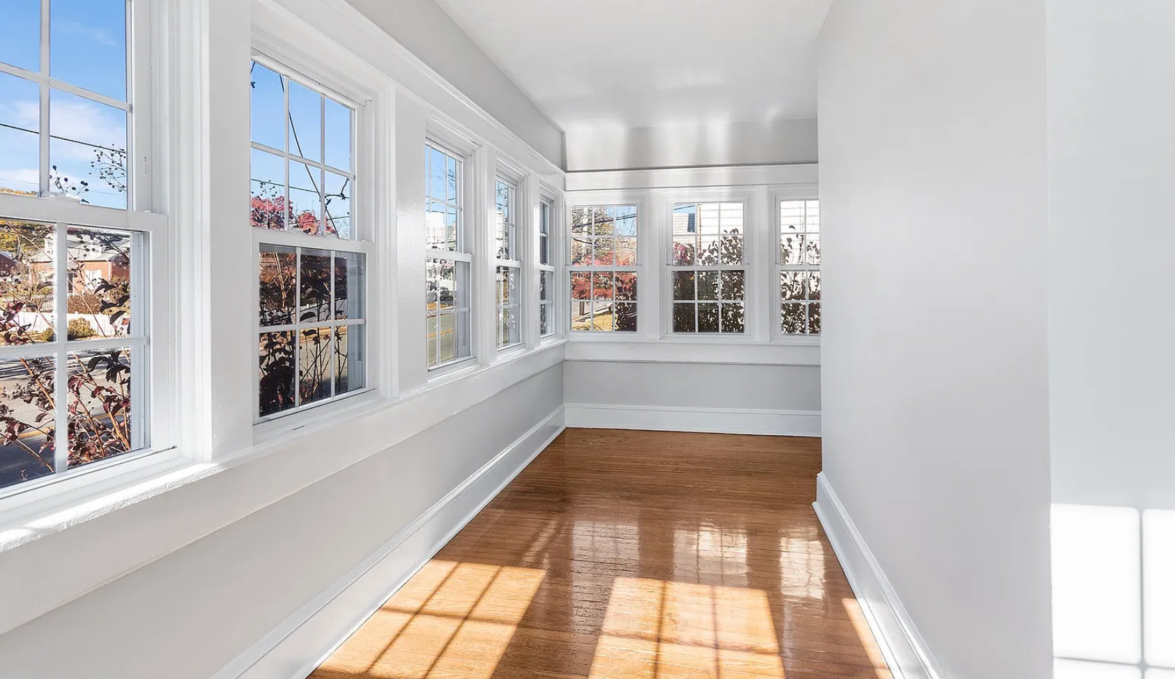 Sunroom with many windows, wooden floor, and white walls. Sunlight streams in.