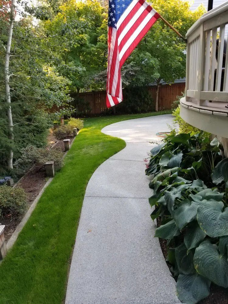 A curving concrete path with green grass on one side, leading towards an American flag and trees.