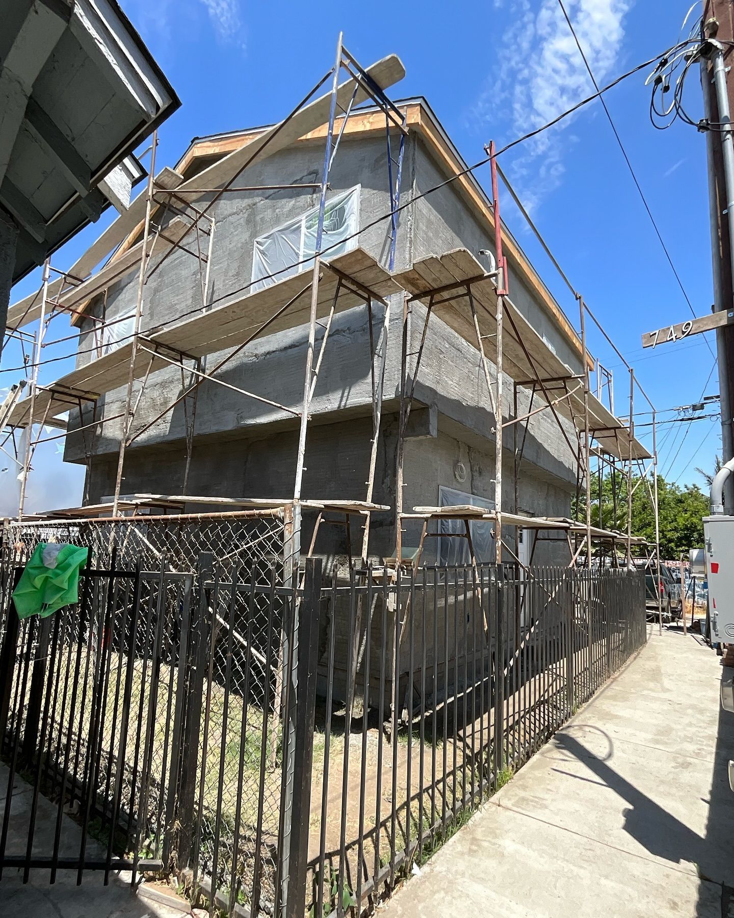 Two-story house under construction with scaffolding, gray stucco, and black fence. Sunny day.