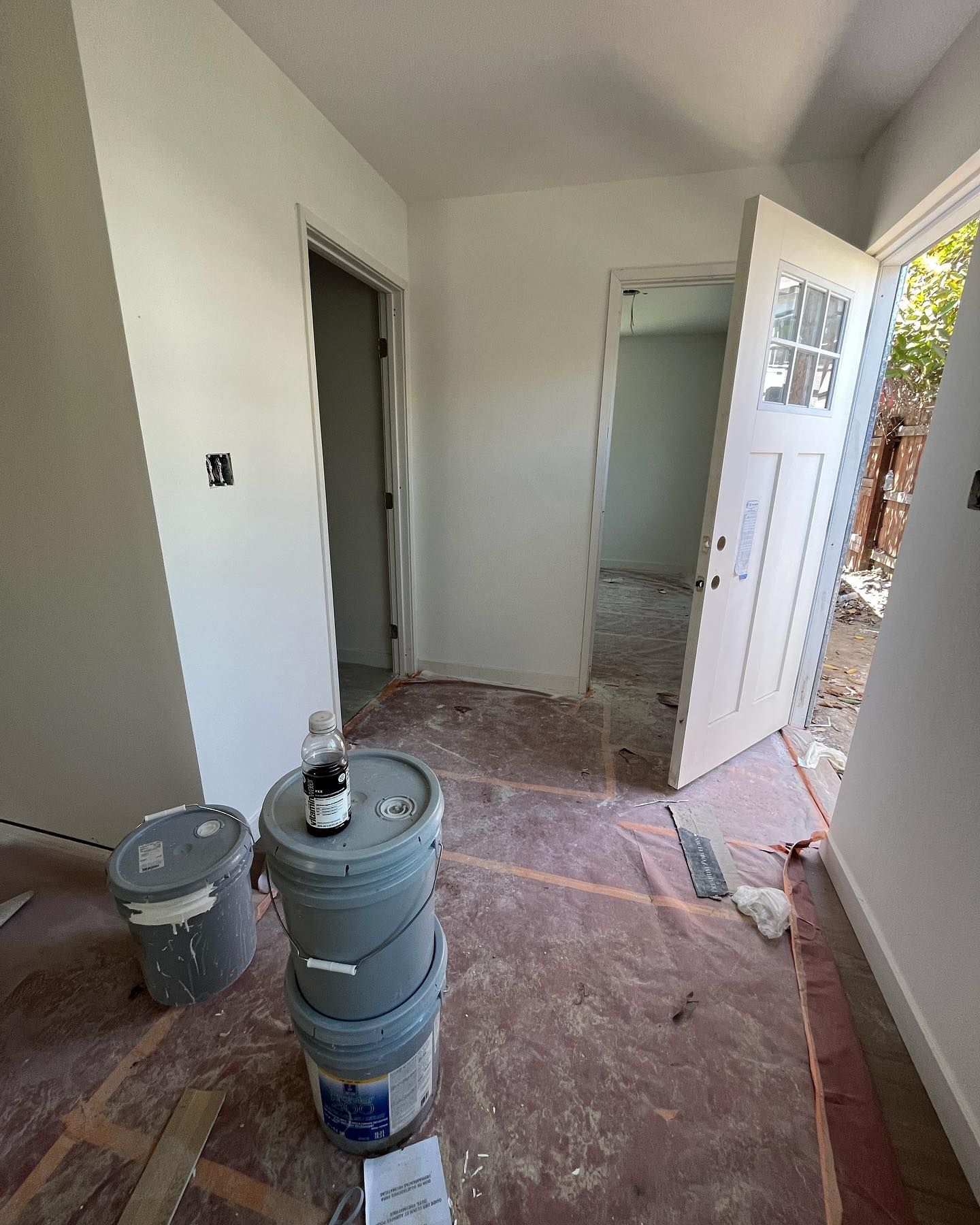 Interior view of a hallway with paint buckets, open doors, and unfinished flooring. Walls are painted white.