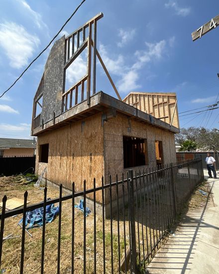A two-story building under construction with exposed wooden framing and plywood walls. A man stands nearby on a sunny day.