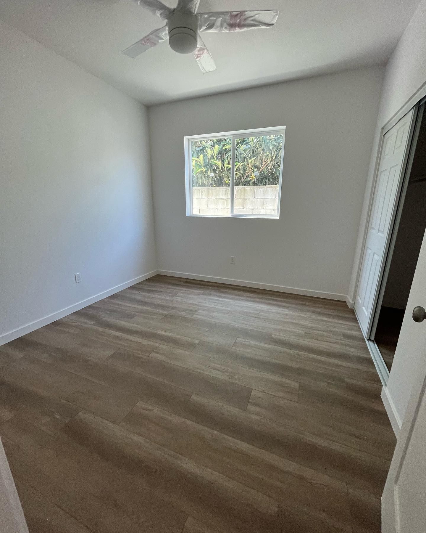 Empty bedroom with wood-look flooring, white walls, a window, sliding closet doors, and a ceiling fan.