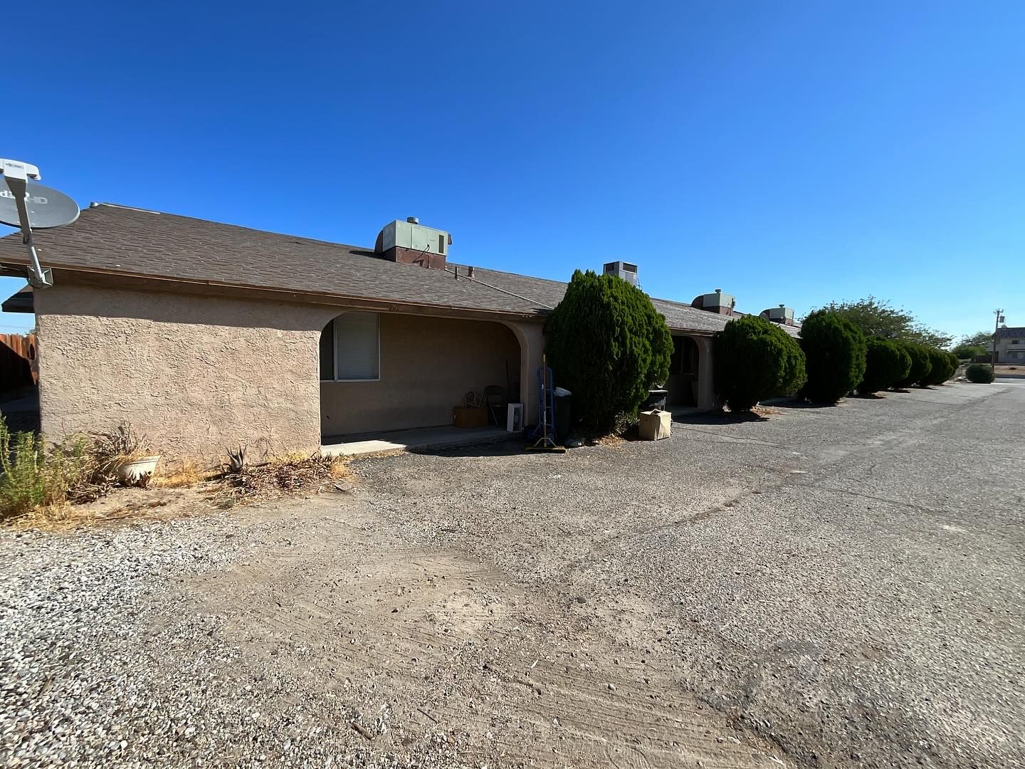 Tan stucco building with a gravel parking area, shrubs, and satellite dishes against a blue sky.