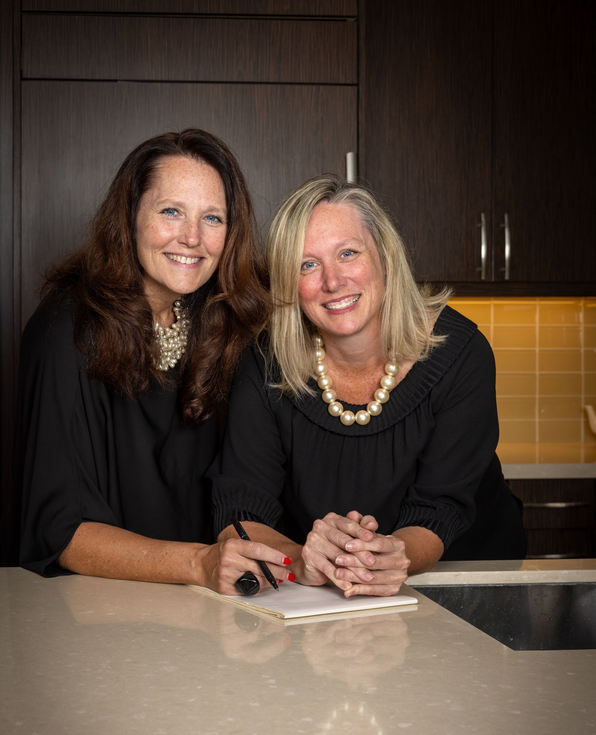 Two women smiling and posing in a kitchen, leaning on a counter.