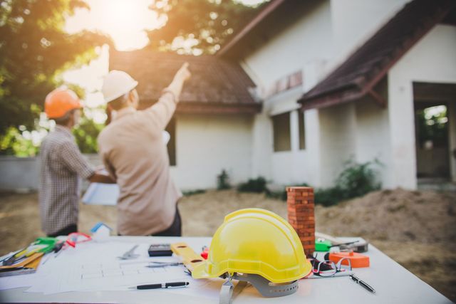 Deux ouvriers du bâtiment, casqués, examinent un plan de maison et désignent la maison du doigt. Des outils sont posés sur la table.